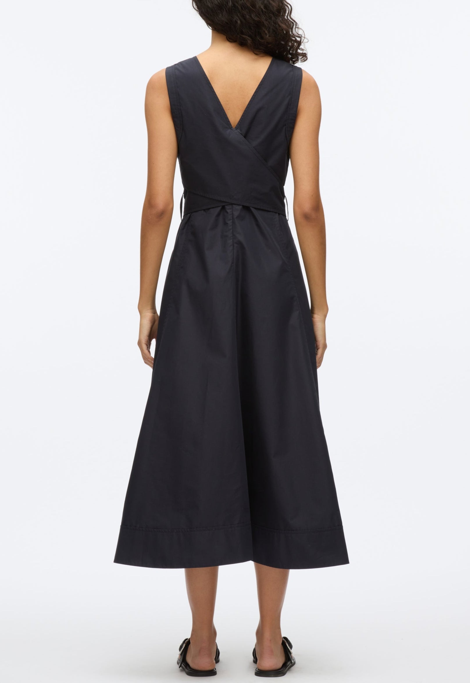 A woman faces away, wearing the 3.1 Phillip Lim Crisscross Back Midi Dress—a sleeveless, dark technical poplin V-neck dress with an A-line skirt—and black shoes, set against a plain white background.