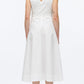 A woman with curly blonde hair models the 3.1 Phillip Lim Crisscross Back Midi Dress—a sleeveless white V-neck A-line dress with a crisscross back—paired with black sandals, shown from behind on a plain white background.