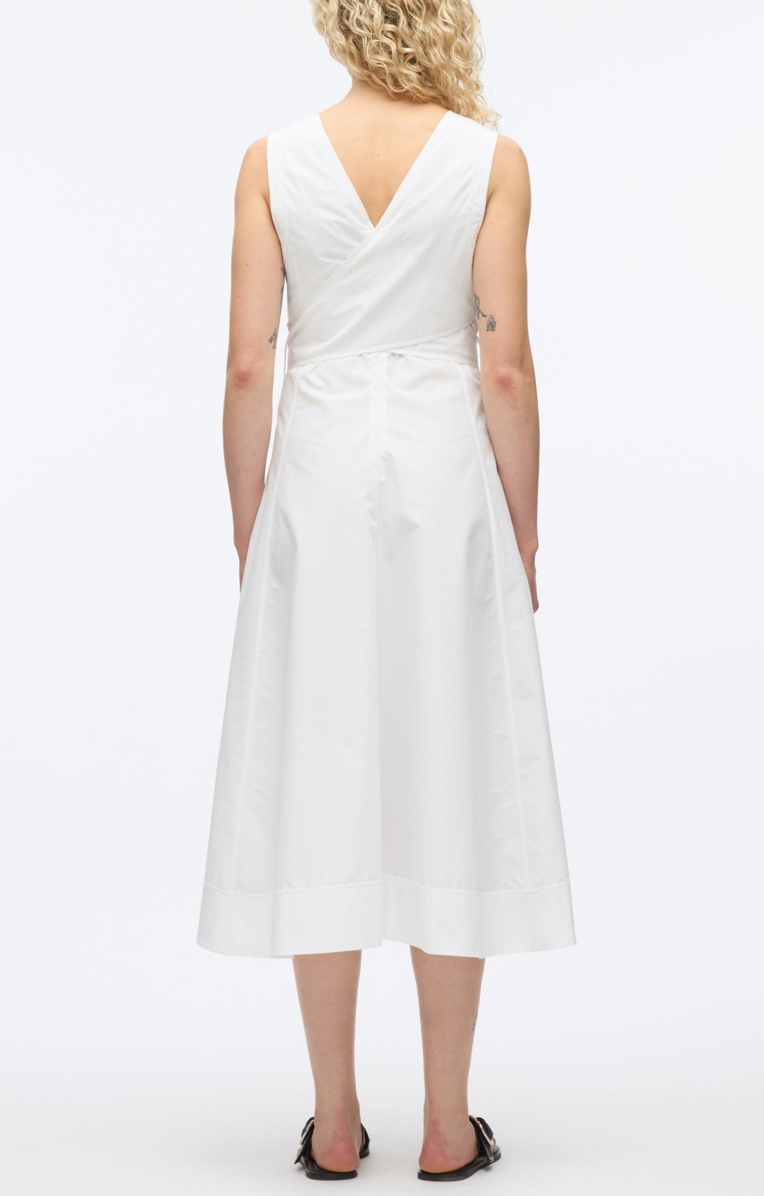 A woman with curly blonde hair models the 3.1 Phillip Lim Crisscross Back Midi Dress—a sleeveless white V-neck A-line dress with a crisscross back—paired with black sandals, shown from behind on a plain white background.