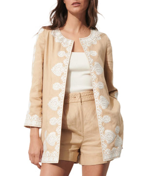 A woman wears the Wren Jacket by Figue—a beige embroidered layer—over a white crew-neck top, paired with matching beige relaxed-fit shorts with a braided waistband.