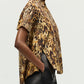 A person poses in profile against a plain background, styled in Adam Lippes’ Charlie Top—short-sleeved, brown and gold with a collared silhouette—paired with black pants.