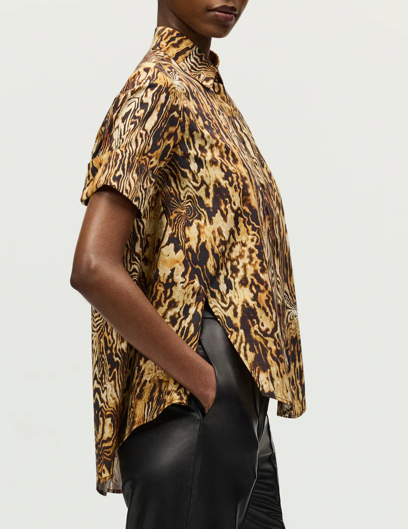 A person poses in profile against a plain background, styled in Adam Lippes’ Charlie Top—short-sleeved, brown and gold with a collared silhouette—paired with black pants.