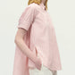 A person models the Adam Lippes Charlie Top—a light pink, short-sleeved, collared shirt in stripe shirting—paired with white pants, shown in profile against a plain background.