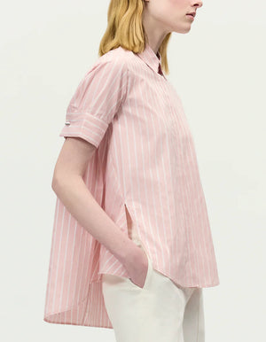 A person models the Adam Lippes Charlie Top—a light pink, short-sleeved, collared shirt in stripe shirting—paired with white pants, shown in profile against a plain background.