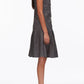 A woman stands in profile wearing the 3.1 Phillip Lim Draped Tank Dress with Floral Applique, a knee-length gray dress featuring a drop waist and pleated skirt, paired with black low-heeled shoes.