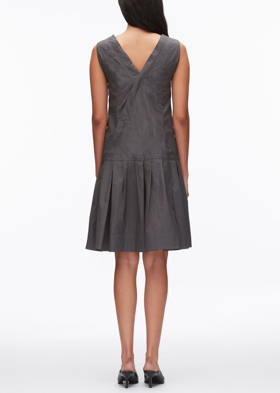 A woman stands facing away, wearing the 3.1 Phillip Lim Draped Tank Dress with Floral Applique—a sleeveless gray knee-length dress featuring a V-shaped back and pleated skirt—paired with black heels.
