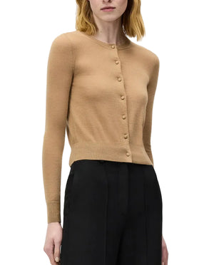 A woman wears the Adam Lippes Worth Cardigan in camel, featuring a classic silhouette and knit-covered buttons, paired with high-waisted black trousers against a plain background.