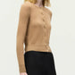 A woman stands against a plain light background wearing the Adam Lippes Worth Cardigan, featuring knit-covered buttons, paired with black high-waisted pants.