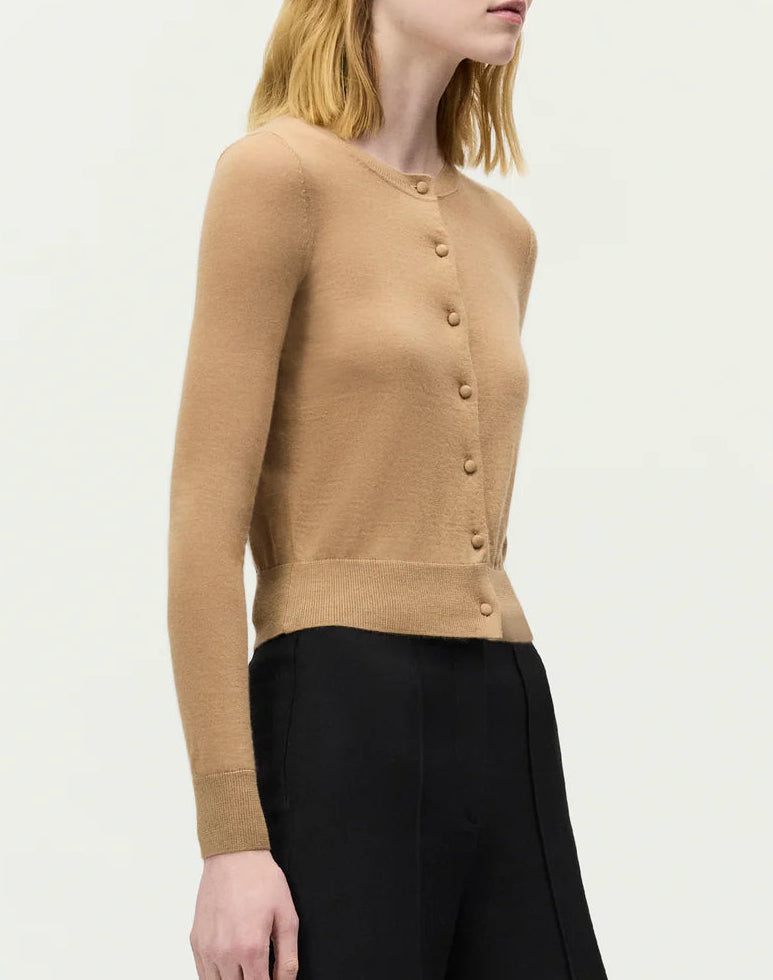 A woman stands against a plain light background wearing the Adam Lippes Worth Cardigan, featuring knit-covered buttons, paired with black high-waisted pants.