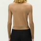 A person with shoulder-length light brown hair is shown from the back against a plain light background, wearing the Adam Lippes Worth Cardigan in tan cashmere-silk with knit-covered buttons and black pants, exuding a classic silhouette.