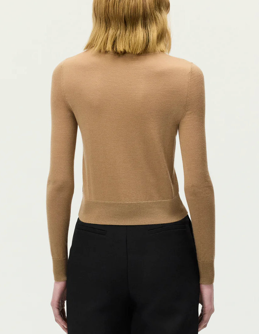 A person with shoulder-length light brown hair is shown from the back against a plain light background, wearing the Adam Lippes Worth Cardigan in tan cashmere-silk with knit-covered buttons and black pants, exuding a classic silhouette.