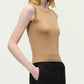 A woman stands against a plain background, wearing the Adam Lippes Ava Top—a sleeveless beige Italian knitwear piece—paired with black trousers, one hand in her pocket.