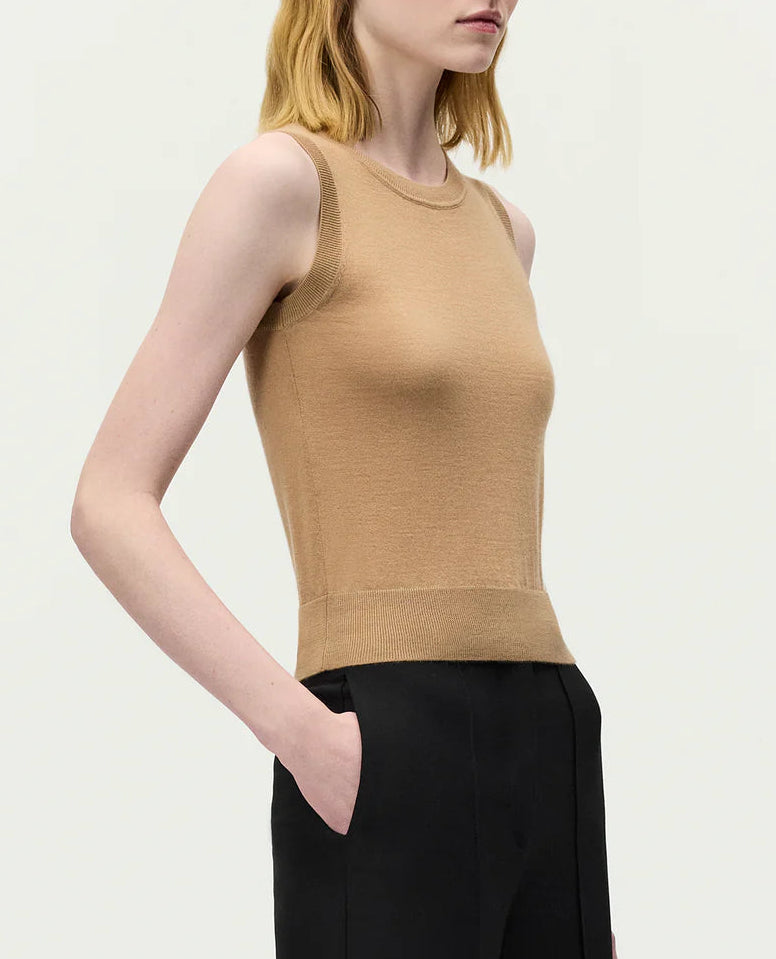 A woman stands against a plain background, wearing the Adam Lippes Ava Top—a sleeveless beige Italian knitwear piece—paired with black trousers, one hand in her pocket.