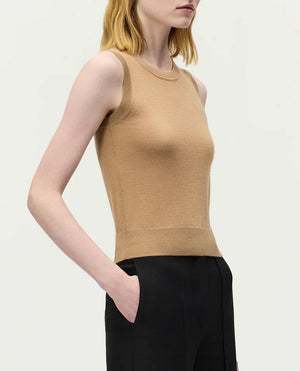 A woman stands against a plain background, wearing the Adam Lippes Ava Top—a sleeveless beige Italian knitwear piece—paired with black trousers, one hand in her pocket.