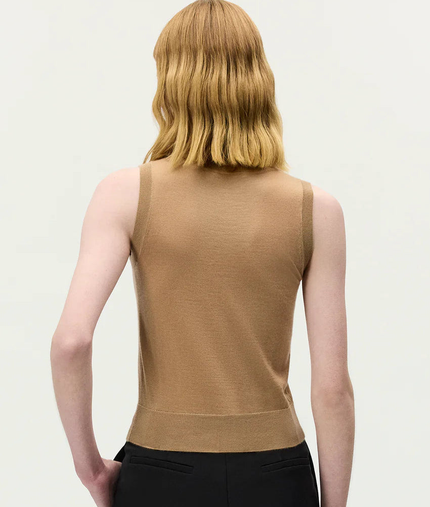 Person with shoulder-length, wavy blonde hair is shown from the back against a plain light background, wearing the Ava Top by Adam Lippes in a sleeveless beige cashmere silk blend and black pants.