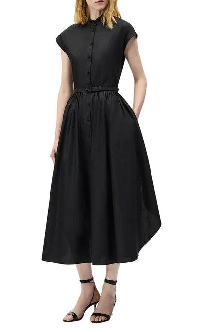A woman in the Adam Lippes Genevieve Dress—a short-sleeve, button-up black shirtdress with a belted waist and pleated skirt—pairs it with black strappy sandals, standing against a white background.