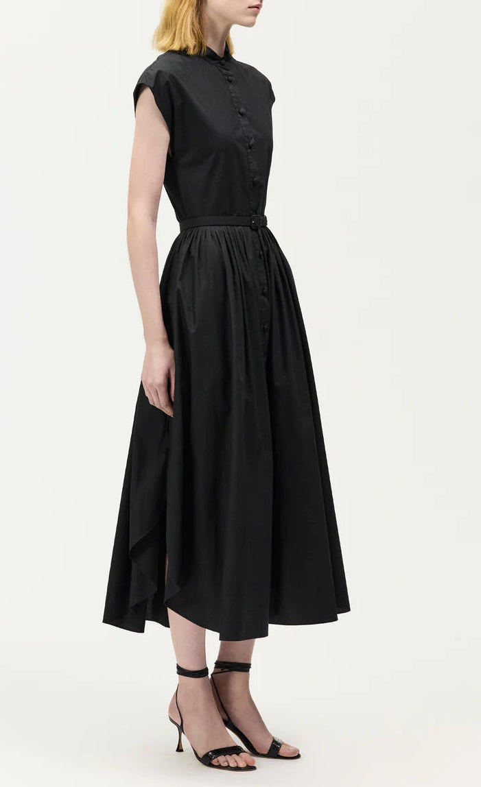 A woman wears the Adam Lippes Genevieve Dress—a sleeveless, button-up black shirtdress with a fitted waist and flared skirt—paired with black high-heeled sandals.