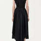 A person stands facing away, wearing the Adam Lippes Genevieve Dress—a black, short-sleeved midi dress with a fitted waist and pleated skirt—paired with black strappy heels.