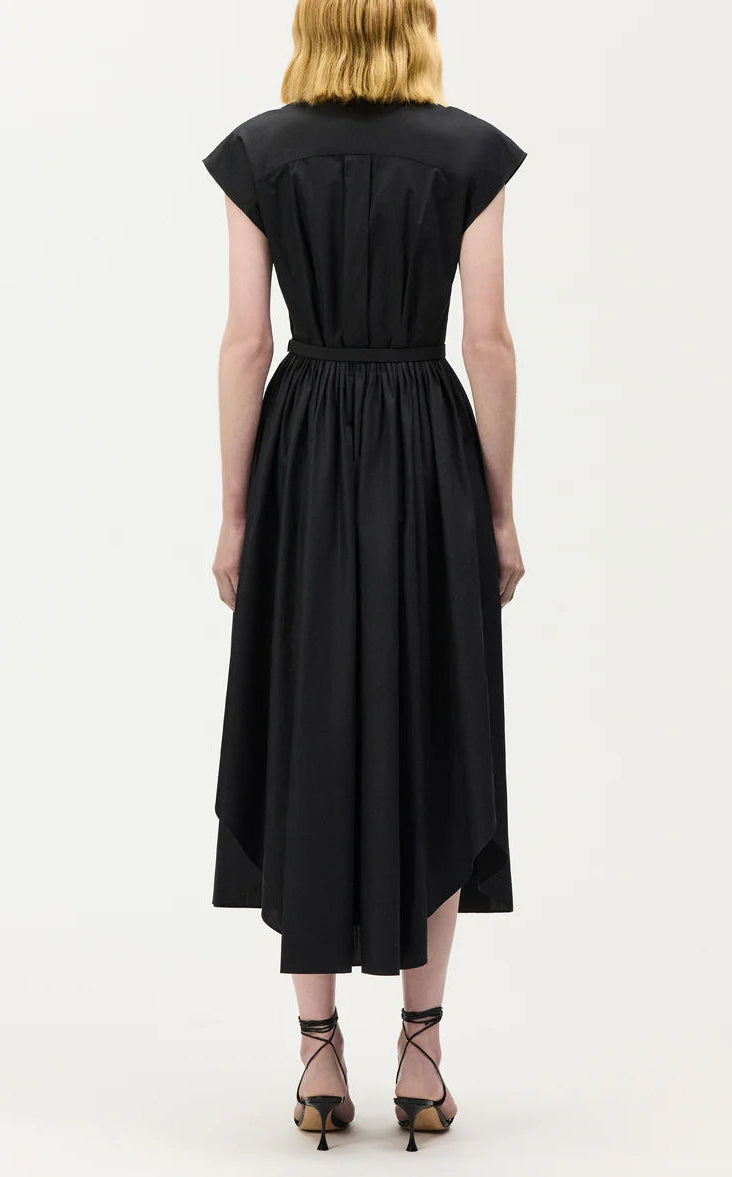 A person stands facing away, wearing the Adam Lippes Genevieve Dress—a black, short-sleeved midi dress with a fitted waist and pleated skirt—paired with black strappy heels.
