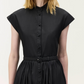 A woman wears the Genevieve Dress by Adam Lippes—a black, short-sleeved shirtdress with a stand collar, belted waist, and pleated skirt for a classic midi look.
