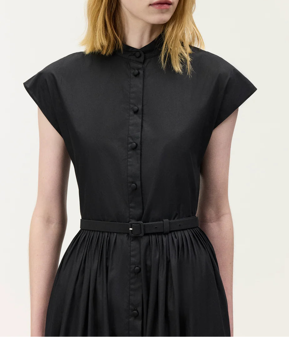 A woman wears the Genevieve Dress by Adam Lippes—a black, short-sleeved shirtdress with a stand collar, belted waist, and pleated skirt for a classic midi look.