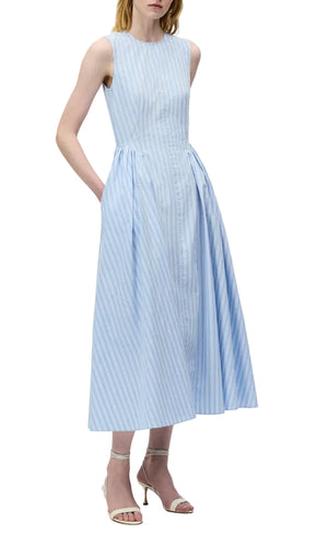 A woman wears the Aliena Dress by Adam Lippes, a sleeveless light blue and white striped fit-and-flare style with a fitted bodice and pleated skirt, styled with strappy heeled sandals.