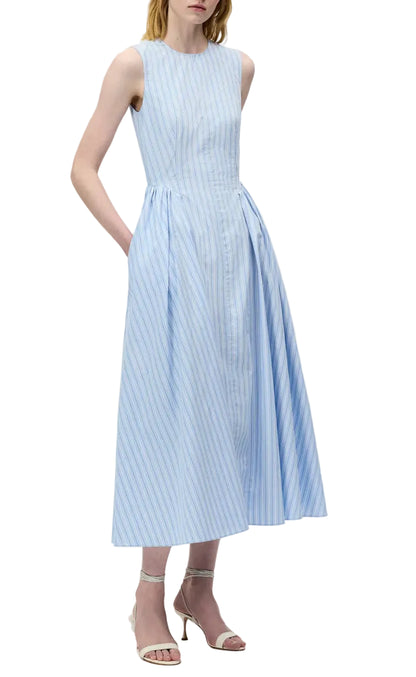 A woman wears the Aliena Dress by Adam Lippes, a sleeveless light blue and white striped fit-and-flare style with a fitted bodice and pleated skirt, styled with strappy heeled sandals.
