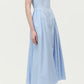 A woman stands against a plain background, wearing the Adam Lippes Aliena Dress—a sleeveless, light blue-striped fit-and-flare style with a fitted bodice and hand-pleated flared skirt—paired with white ankle-strap heels.