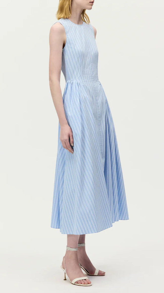 A woman stands against a plain background, wearing the Adam Lippes Aliena Dress—a sleeveless, light blue-striped fit-and-flare style with a fitted bodice and hand-pleated flared skirt—paired with white ankle-strap heels.