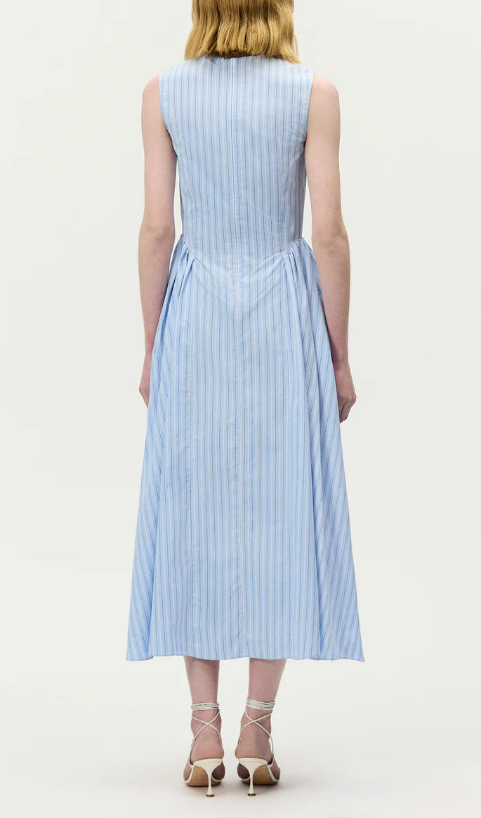A person stands facing away, wearing the Adam Lippes Aliena Dress—a sleeveless, light blue and white vertical striped shirtdress—with strappy heels against a plain background.