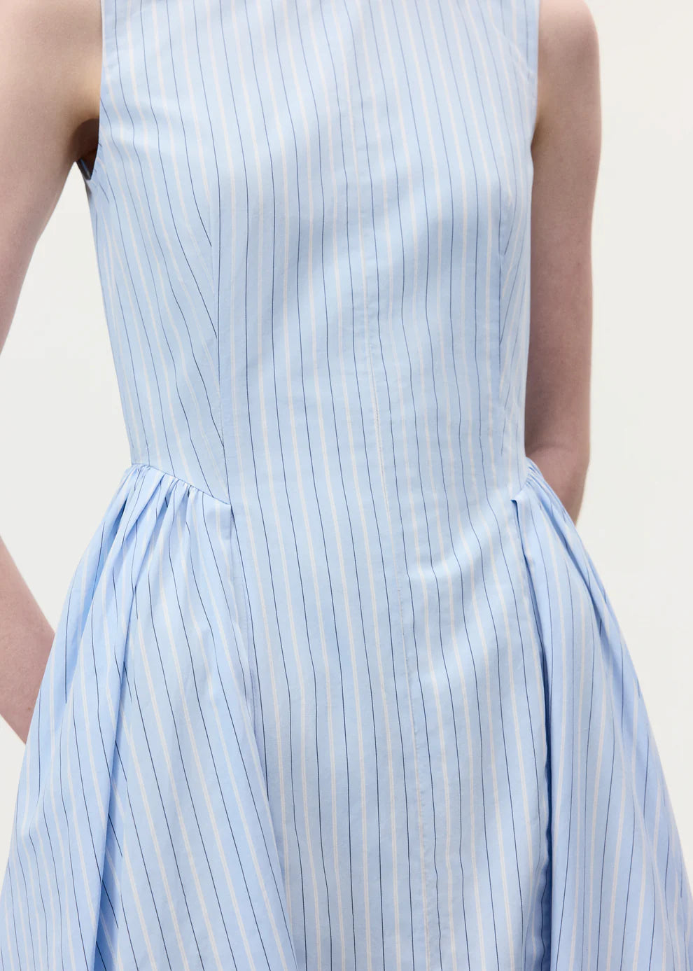 A person wearing the Aliena Dress by Adam Lippes, a sleeveless light blue shirtdress with thin vertical white and dark blue stripes, shown from shoulders to hips.
