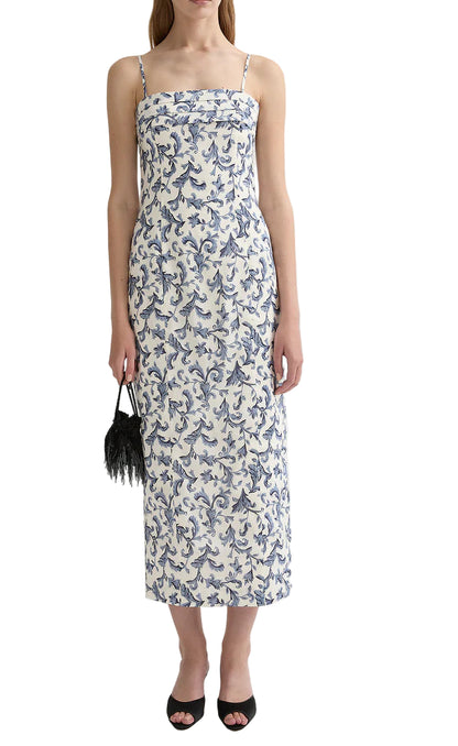 A woman wears the POSSE Rue Dress, a white linen silk dress with blue floral print, thin straps, and square neckline, paired with a black feathered clutch and black open-toe heels.