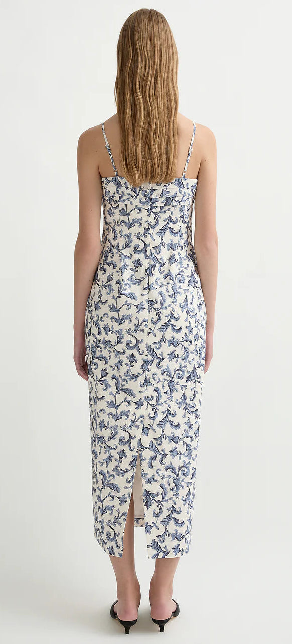 A woman with long, wavy hair is seen from the back wearing the POSSE Rue Dress—an ankle-length, sleeveless linen silk dress with blue floral patterns, a fitted silhouette, back slit, and paired with black sandals.
