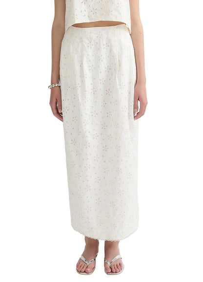 A person wearing the POSSE Salma Column Skirt, a white high-rise embroidered cotton piece, with a matching sleeveless top, silver sandals, and a bracelet.
