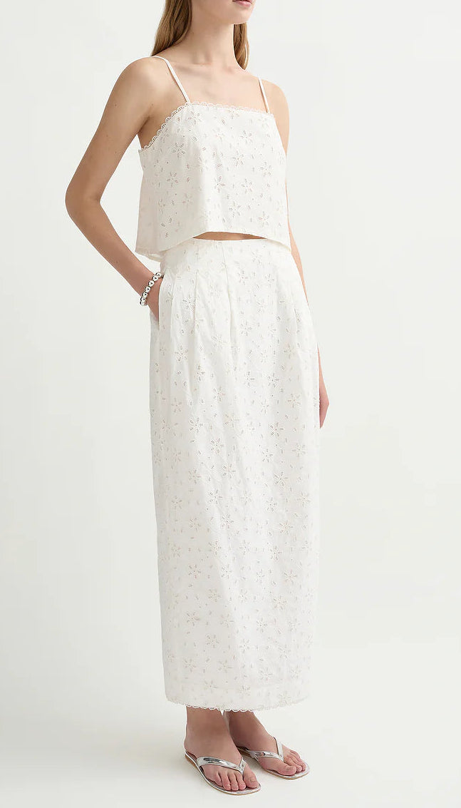 A woman wearing POSSE’s Salma Column Skirt—an embroidered white high-rise cotton skirt with eyelet pattern—paired with a matching sleeveless crop top and flip-flop sandals.