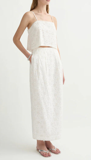 A woman wearing POSSE’s Salma Column Skirt—an embroidered white high-rise cotton skirt with eyelet pattern—paired with a matching sleeveless crop top and flip-flop sandals.