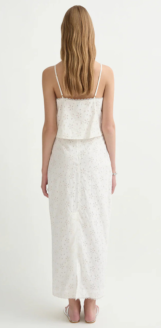 A woman with long wavy hair faces away, wearing a white sleeveless top and the POSSE Salma Column Skirt, paired with flat sandals against a plain background.