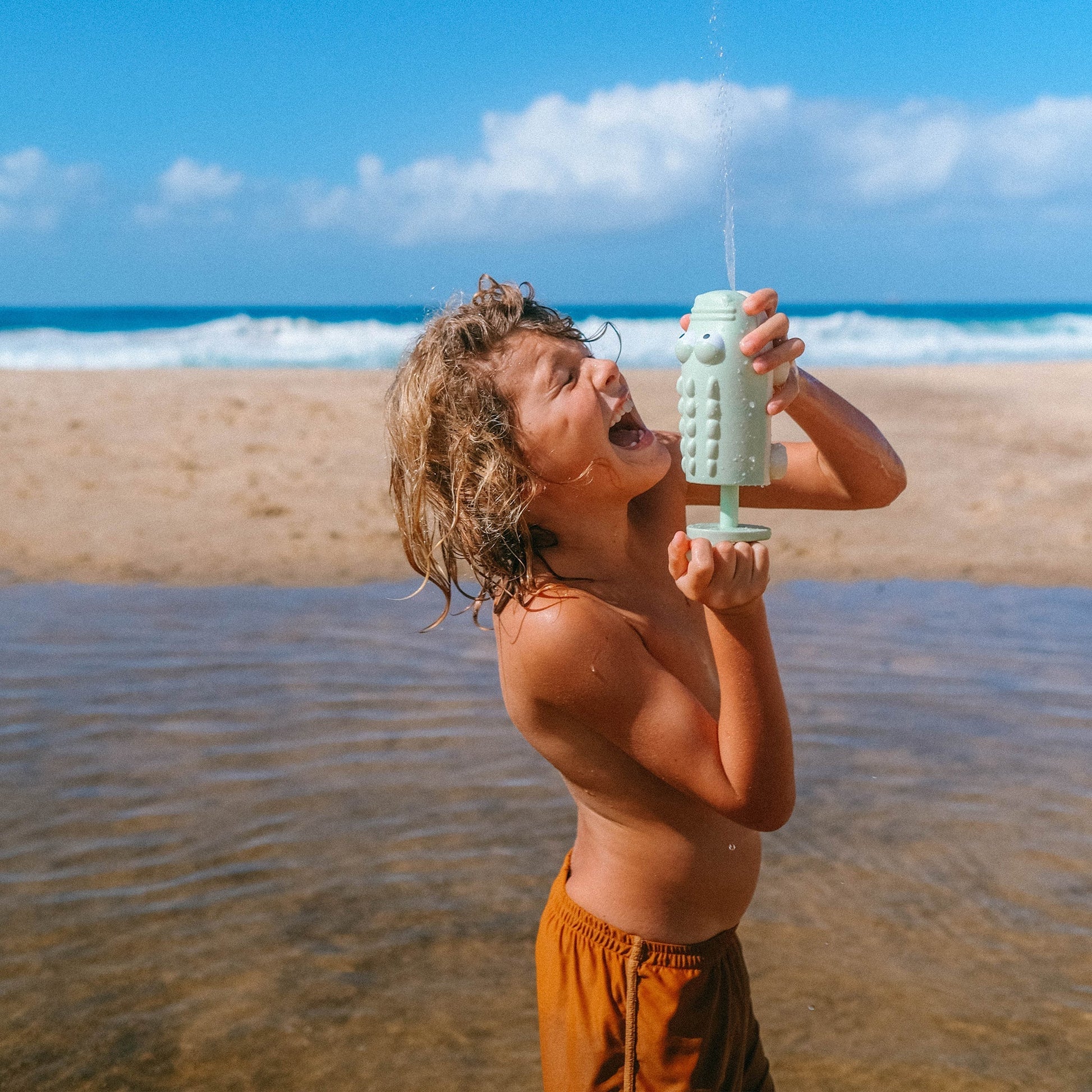 A young boy laughing as he squirts water into his mouth from a Sunnylife Water Squirters at the beach.