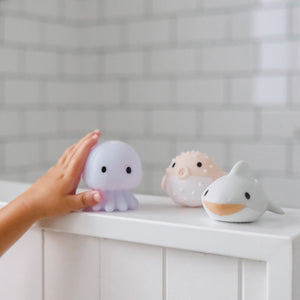 A child’s hand reaches for a light-up octopus bath toy next to Sunnylife Sea Animal Splash Toys shaped like a pufferfish and shark on a tub ledge.