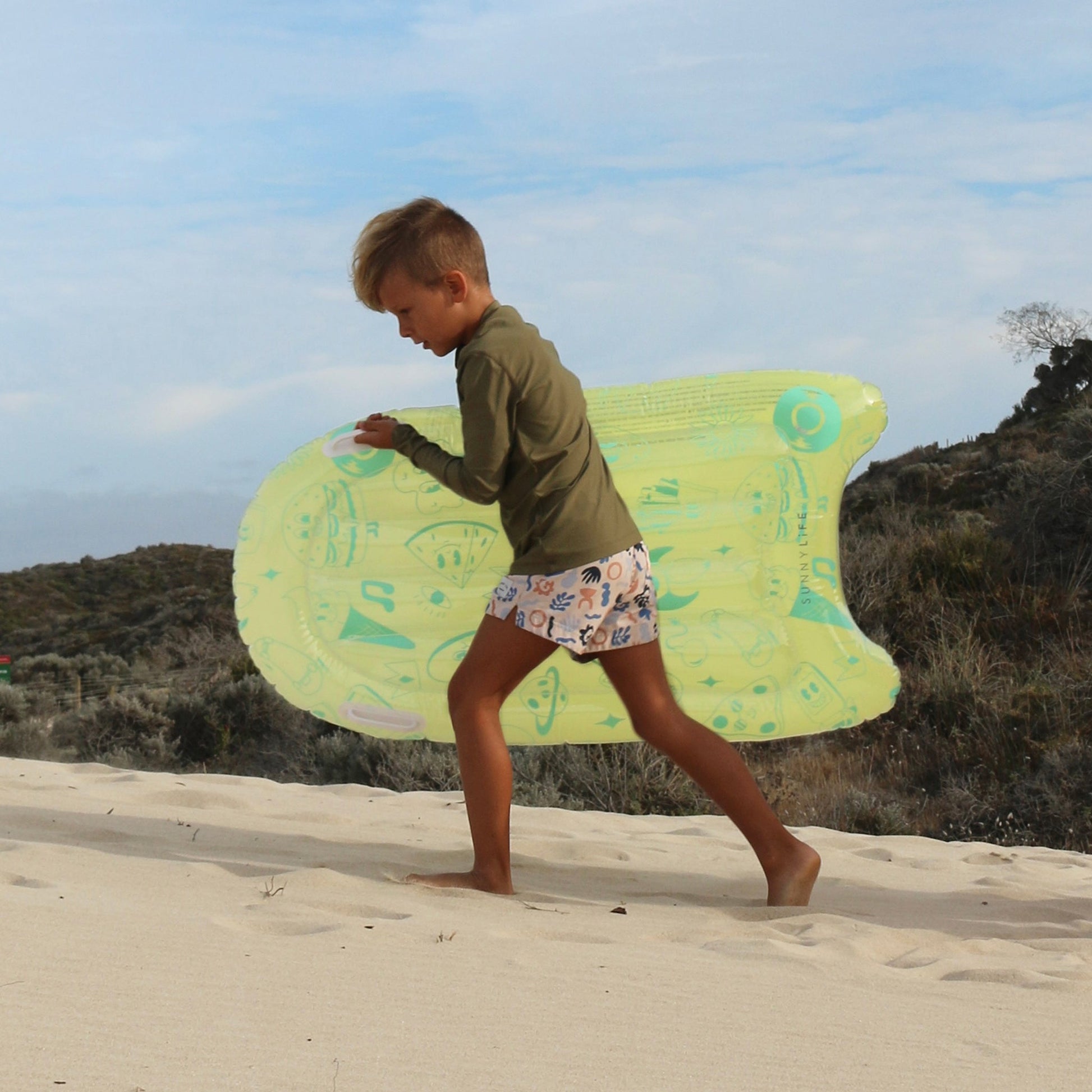 A child walks across sandy terrain carrying a Sunnylife Inflatable Boogie Board by Sunnylife. The child wears a long-sleeve green shirt and white shorts, with a non-vegetated hilly landscape in the background.