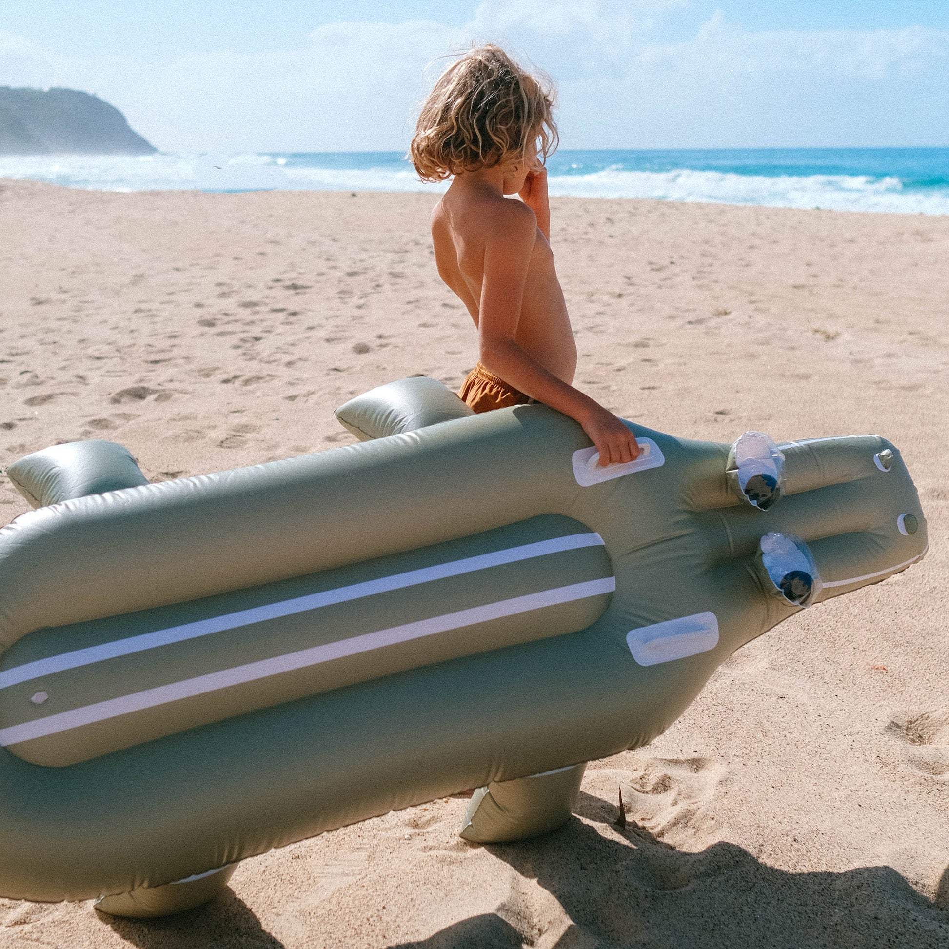 A child stands on a sandy beach holding a large Sunnylife Lie-On Float, Cookie the Croc Khaki, with the ocean and distant cliffs in the background.