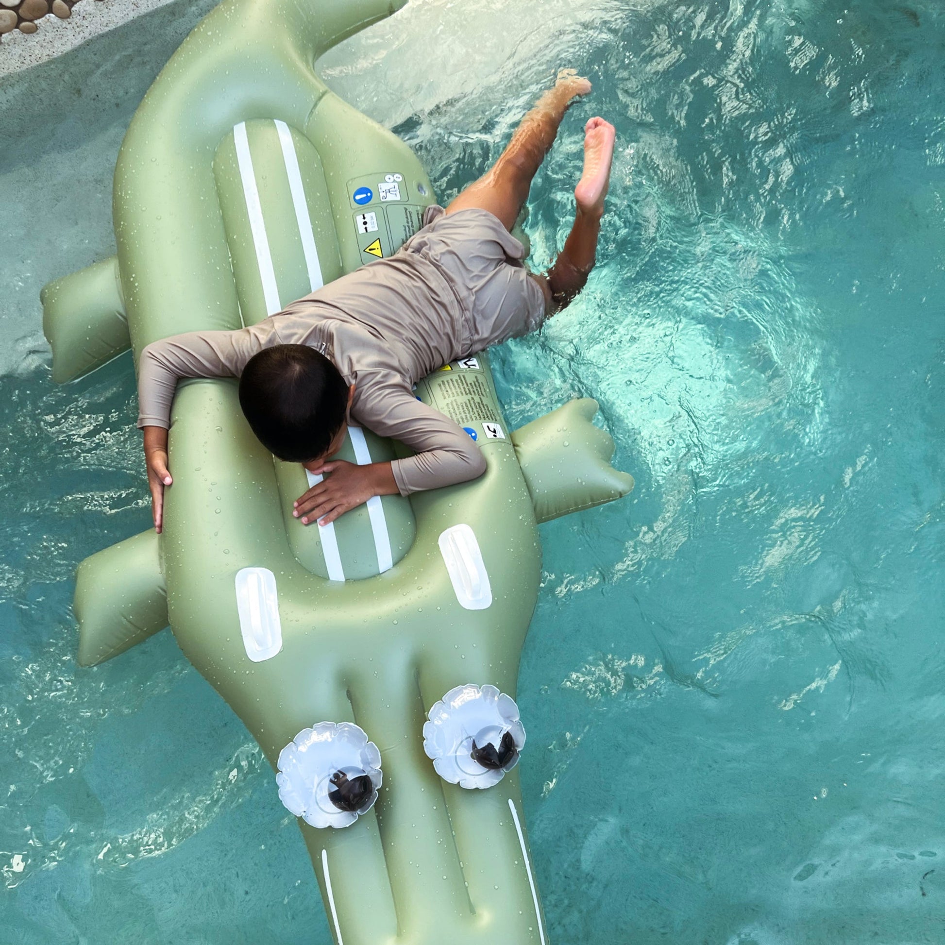 A child in a swimsuit lies face down on the Sunnylife Lie-On Float, Cookie the Croc Khaki, ready for a poolside adventure in the sparkling swimming pool.