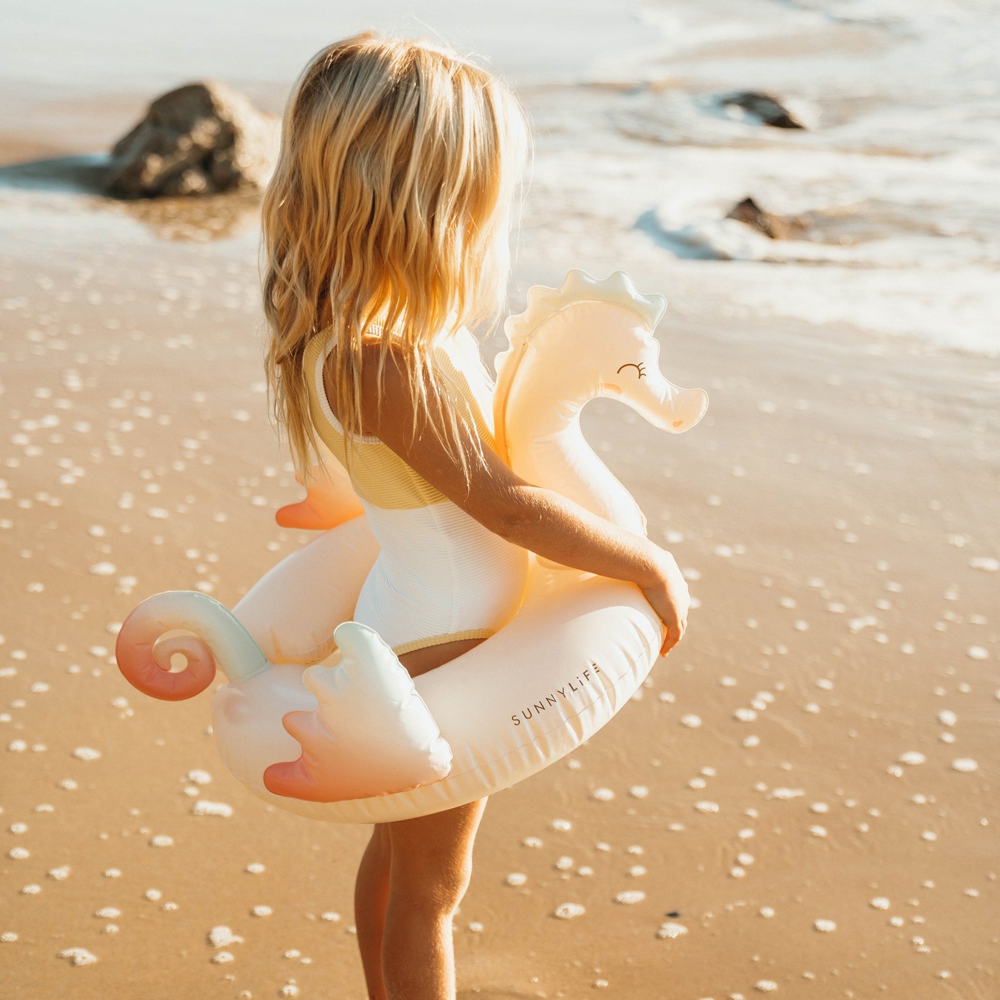 A girl in a swimsuit on a beach with a non-toxic Phthalate free PVC Sunnylife Kids Tube Pool Ring, Melody the Mermaid.