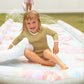 A child with wet hair in a green swimsuit sits on an inflatable water slide, smiling and splashing water, while another child enjoys the Sunnylife Slip Slide Boogie Board Set, Tie Dye in the background.