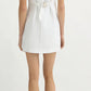 A woman faces away, wearing the POSSE Emma Mini Dress—a sleeveless white linen mini dress with an adjustable back buckle and belt detail—paired with open-toe shoes, set against a plain light background.