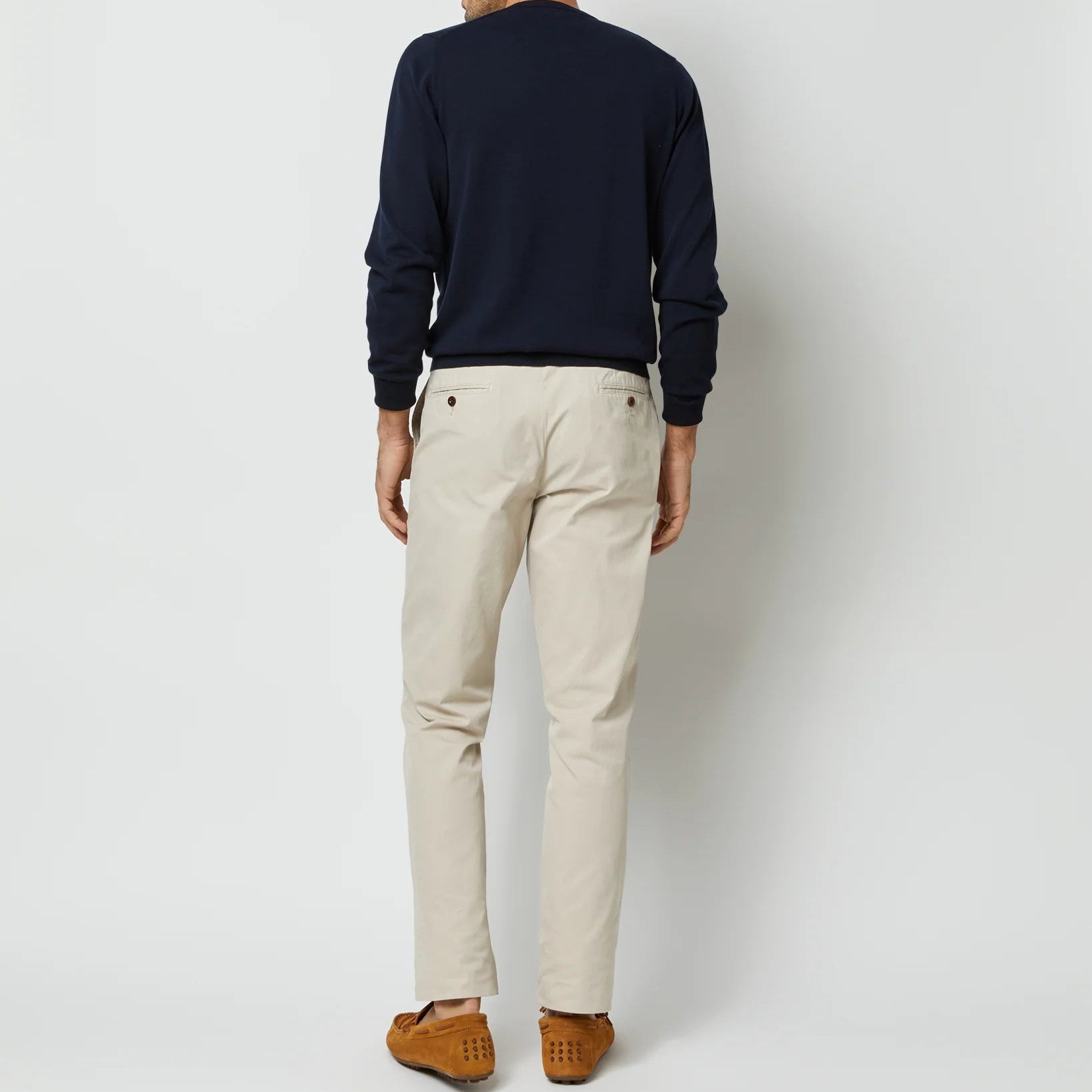 A person stands facing away, wearing a Sid Mashburn Crewneck Sweater in navy, beige pants, and brown loafers against a plain light background.