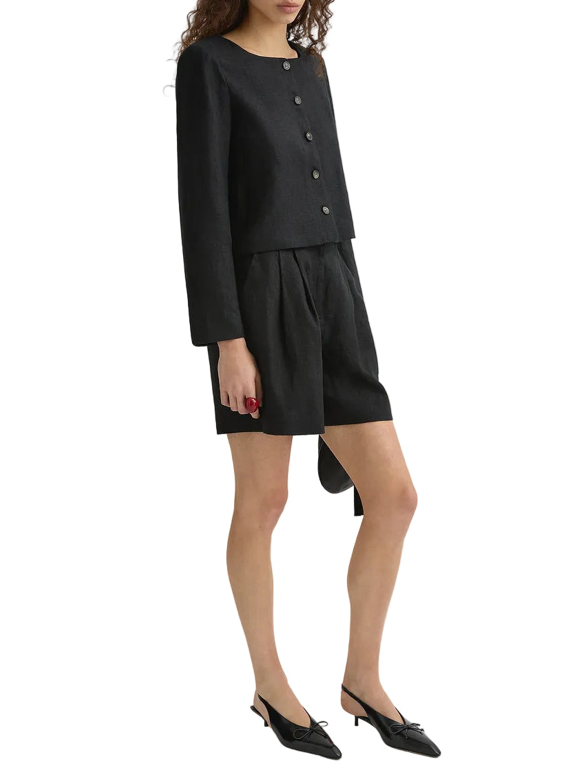 A woman stands against a plain white background wearing the POSSE Marchello Short with a black long-sleeve buttoned top and black low-heeled shoes.