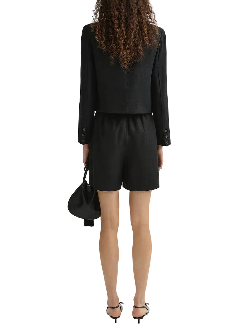A person with curly hair is seen from behind, wearing a black blazer, POSSE’s Marchello Short, heeled sandals, and holding a black handbag.