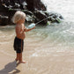 A child with blonde hair, wearing black shorts, joyfully plays at the beach with a SUNNYLiFE's Animal Soaker Shark Pastel Blue water gun, aiming at the water. Waves and rocks create a picturesque backdrop for this delightful scene.