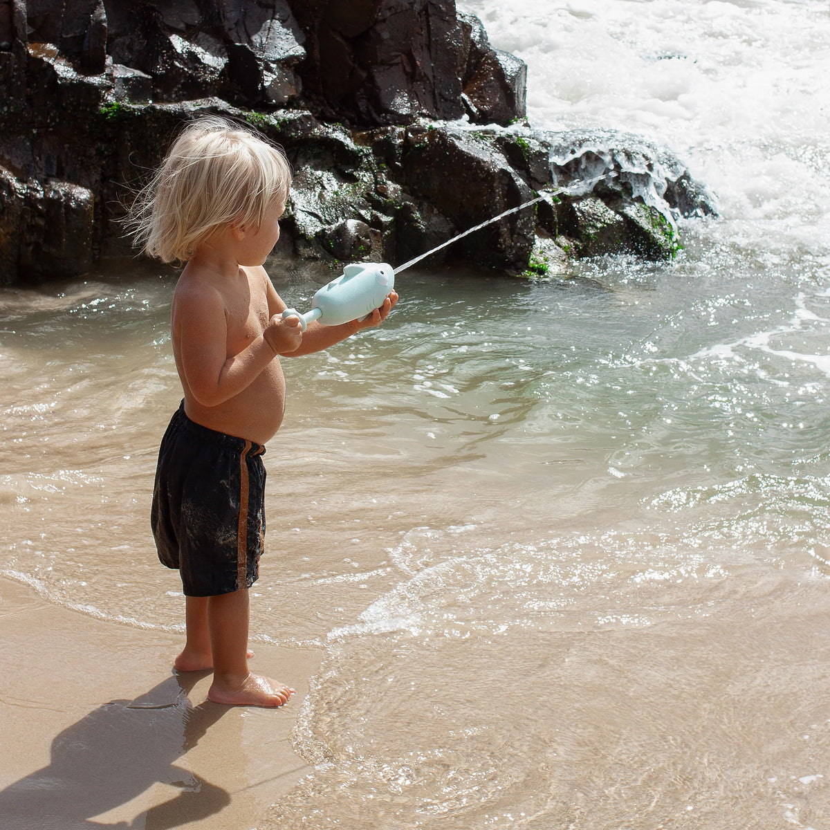 A child with blonde hair, wearing black shorts, joyfully plays at the beach with a SUNNYLiFE's Animal Soaker Shark Pastel Blue water gun, aiming at the water. Waves and rocks create a picturesque backdrop for this delightful scene.